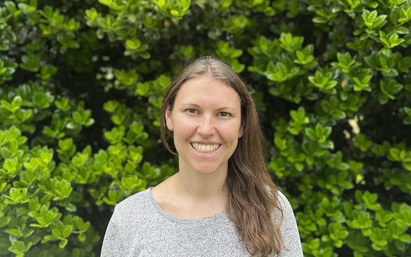 Elizabeth Beu Bourgeois wearing a grey shirt and glasses in front of a vine covered wooden fence